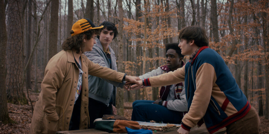 A photo of Gaten Matarazzo, Noah Schnapp, Caleb McLaughlin and Finn Wolfhard standing up and facing each other around a picnic table in a woodsy setting. It is late fall and the leaves in the trees behind them are yellow and sparse. They have their hands together over the picnic table and are looking at each other with resigned yet hopeful faces. They are dressed for fall in medium weight jackets