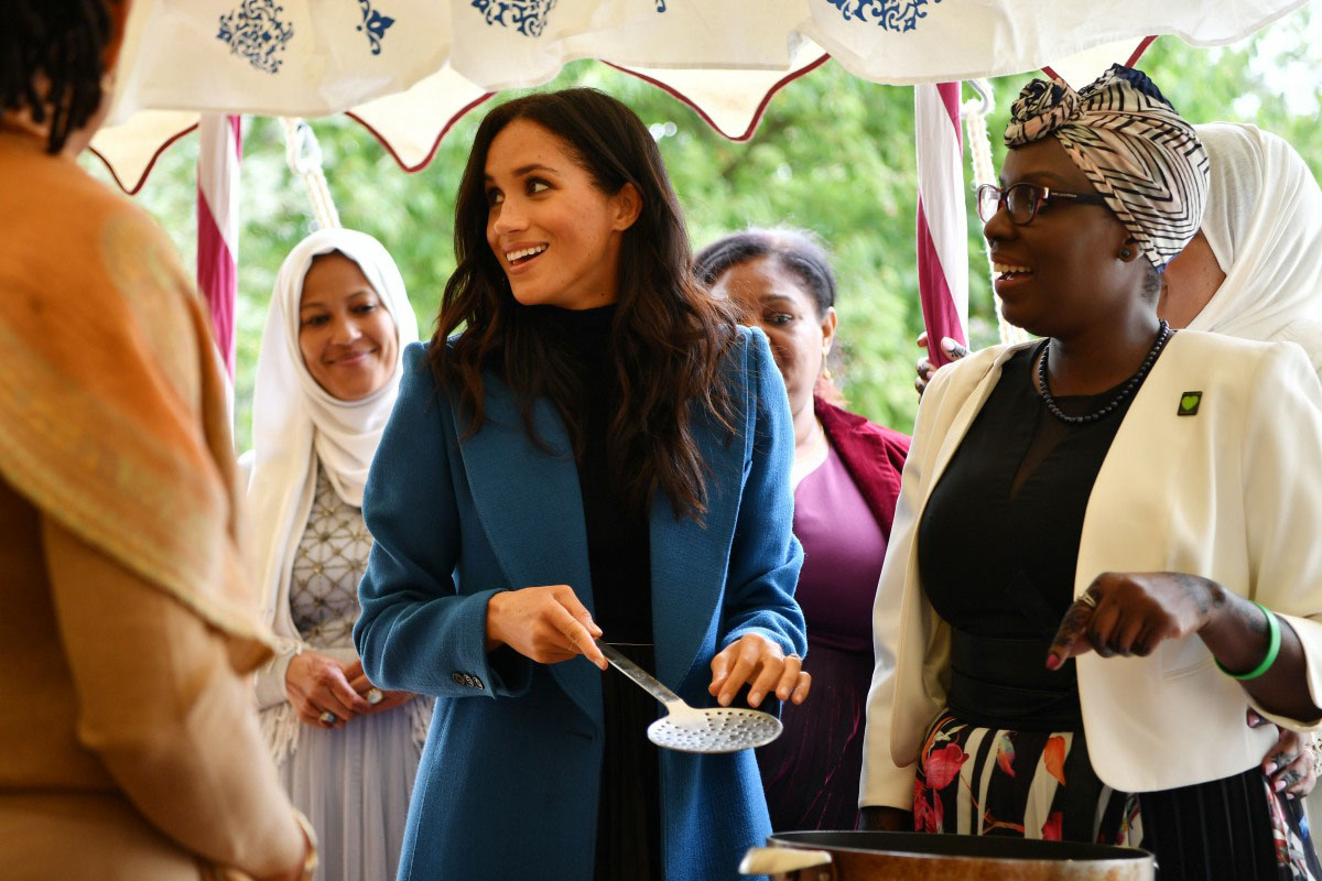 Duchess Meghan at an event for Hubb Community Kitchen. Her head is turned to the side, she is smiling and holding a spoon
