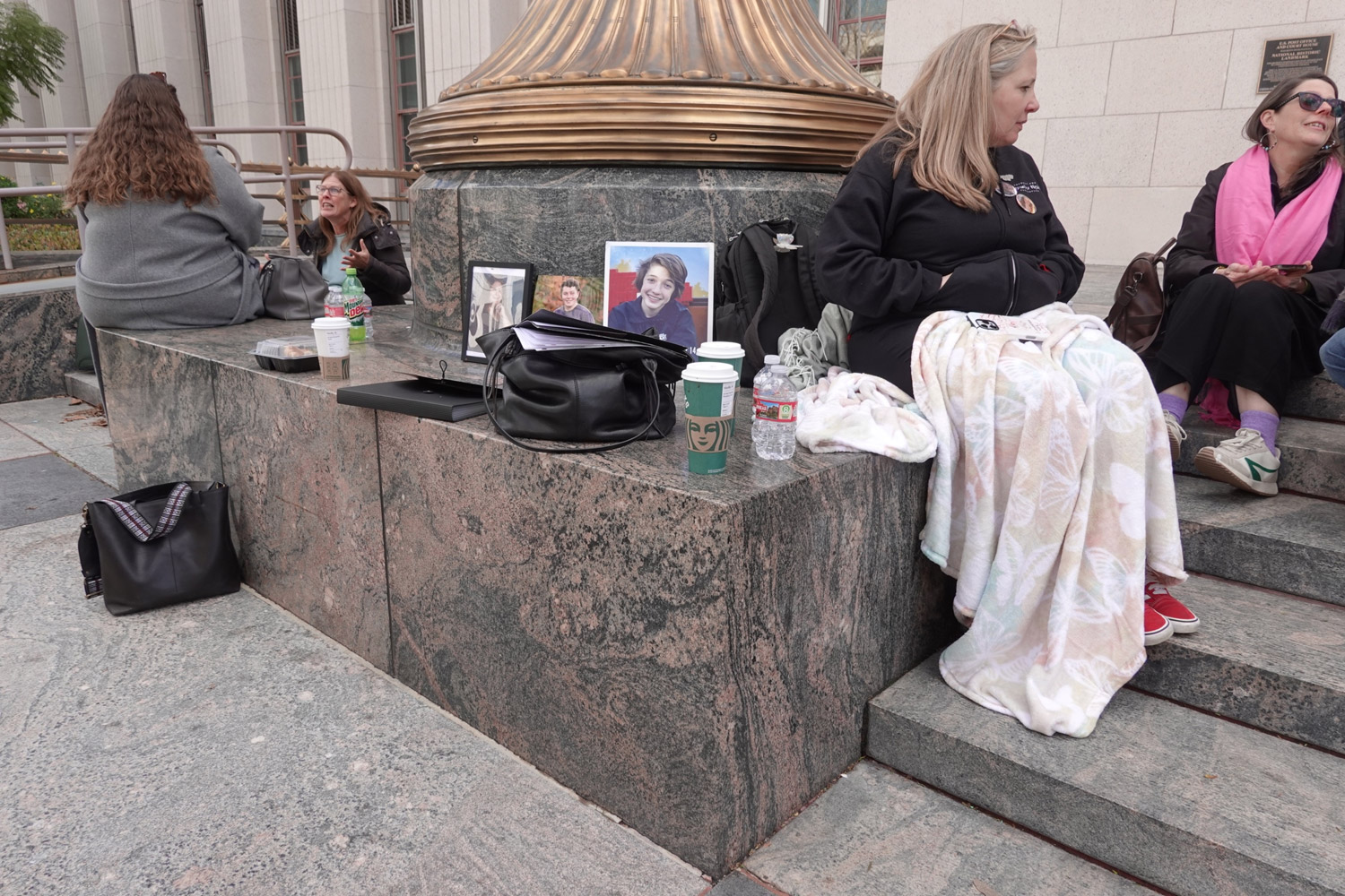 Woman sits in blanket on ledge outside LA court on 2-18-26. Next to her are photos of teens who died after Facebook use