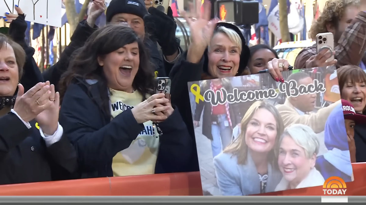 A woman in the crowd outside Today holds a Welcome Back sign with a photo of herself and Savannah and a yellow ribbon on it