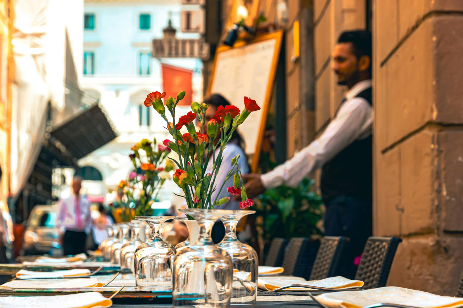 An outdoor restaurant in Italy with floral centerpiece, empty glassware and waiter in the background