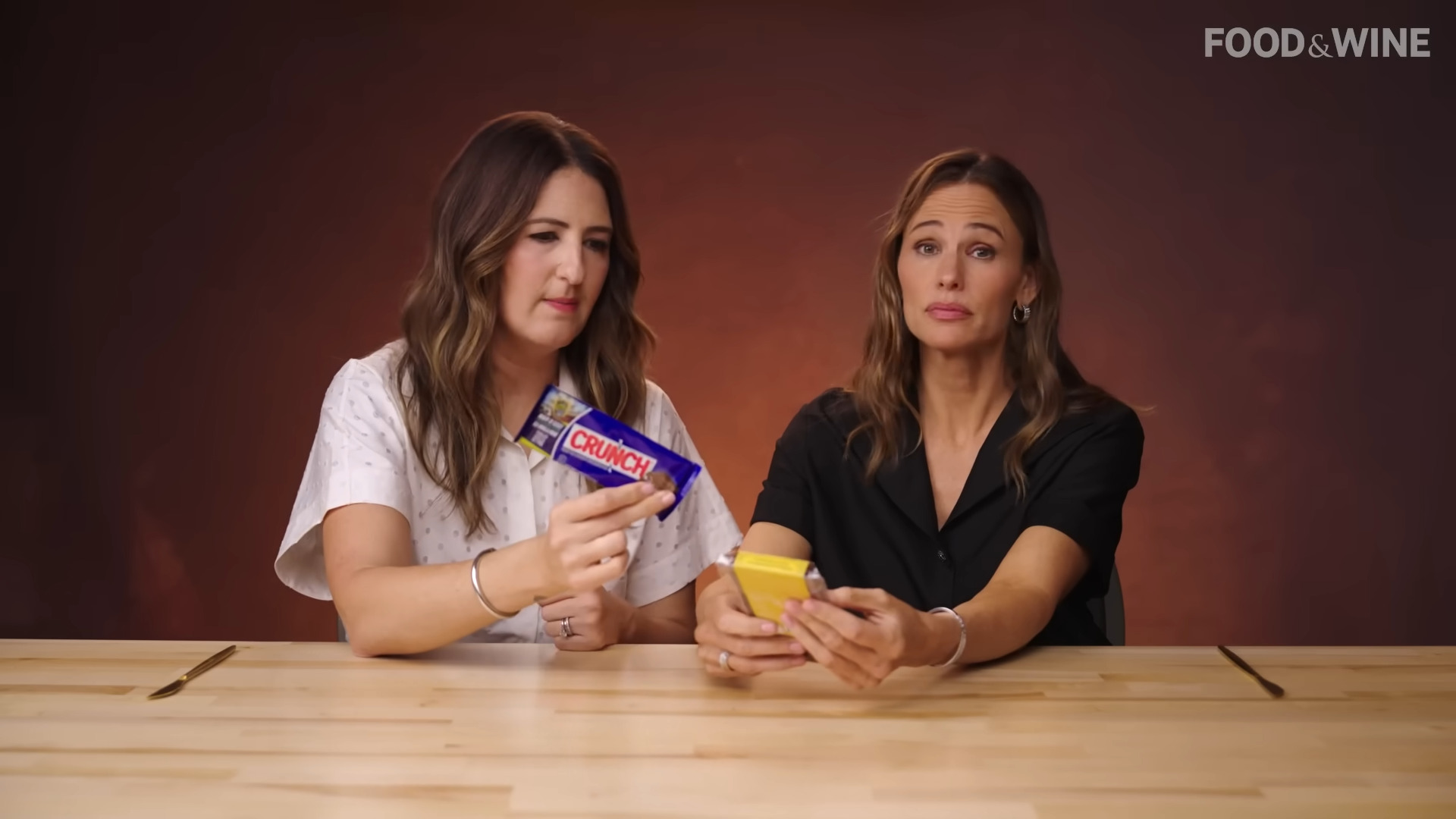 D'arcy Carden and Jennifer Garner holding up Nestle Crunch and Dubai Chocolate Bars