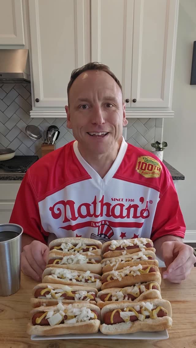 Screenshot of Joey Chestnut with Nathan's shirt and a bunch of hotdogs in front of him in a kitchen
