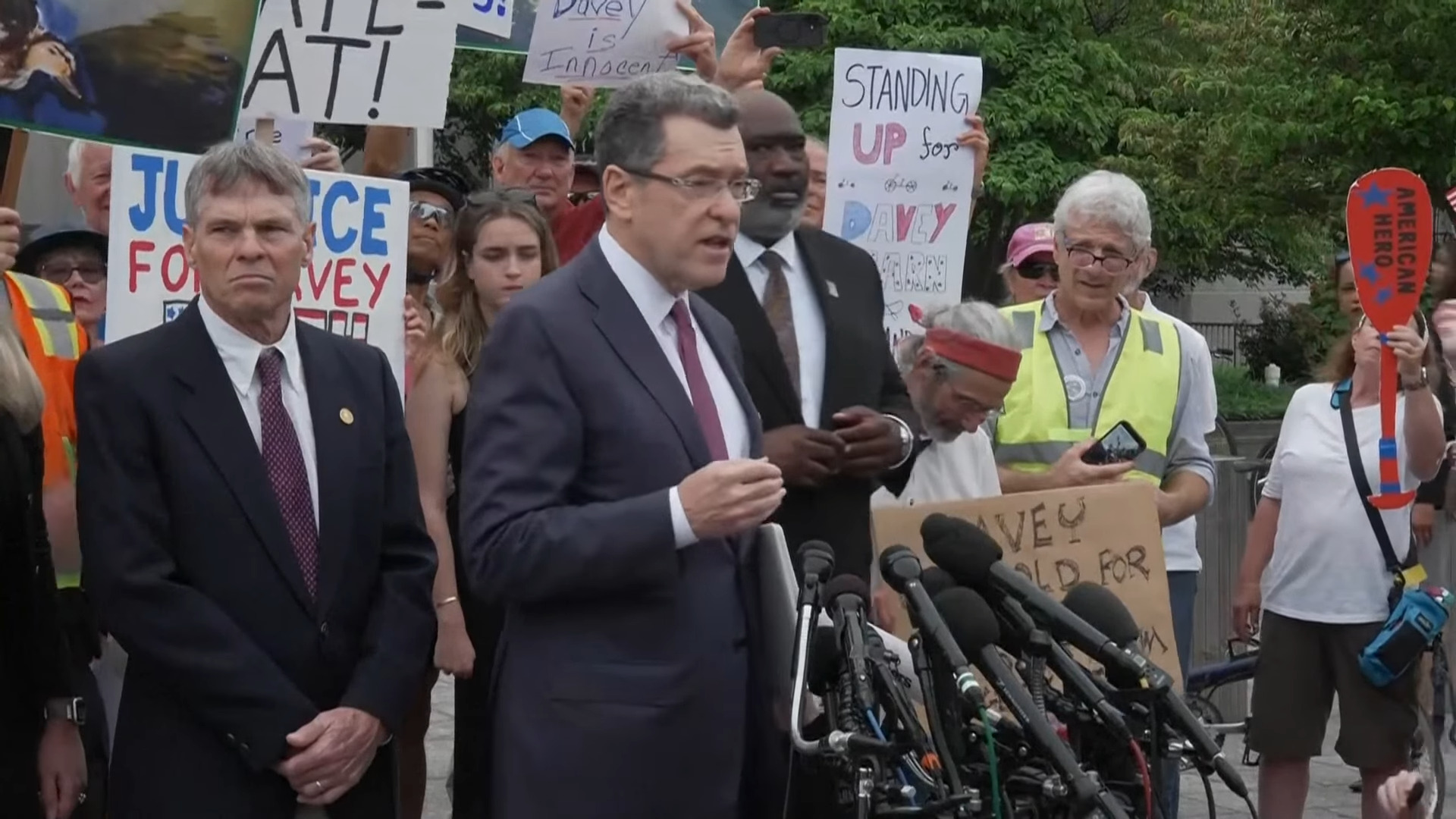 Screenshot of David Hearn and his lawyer speaking to the press outside court. Multiple supportive protestors him are behind him