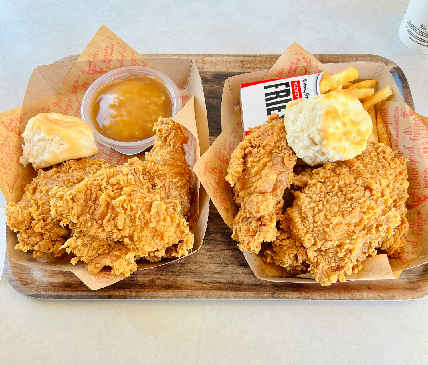 two serving baskets of fried chicken on trays with biscuits and sides