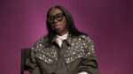 "Person with long hair and glasses, wearing a studded olive jacket and white shirt, sits on a chair against a plain purple background in a studio setting."